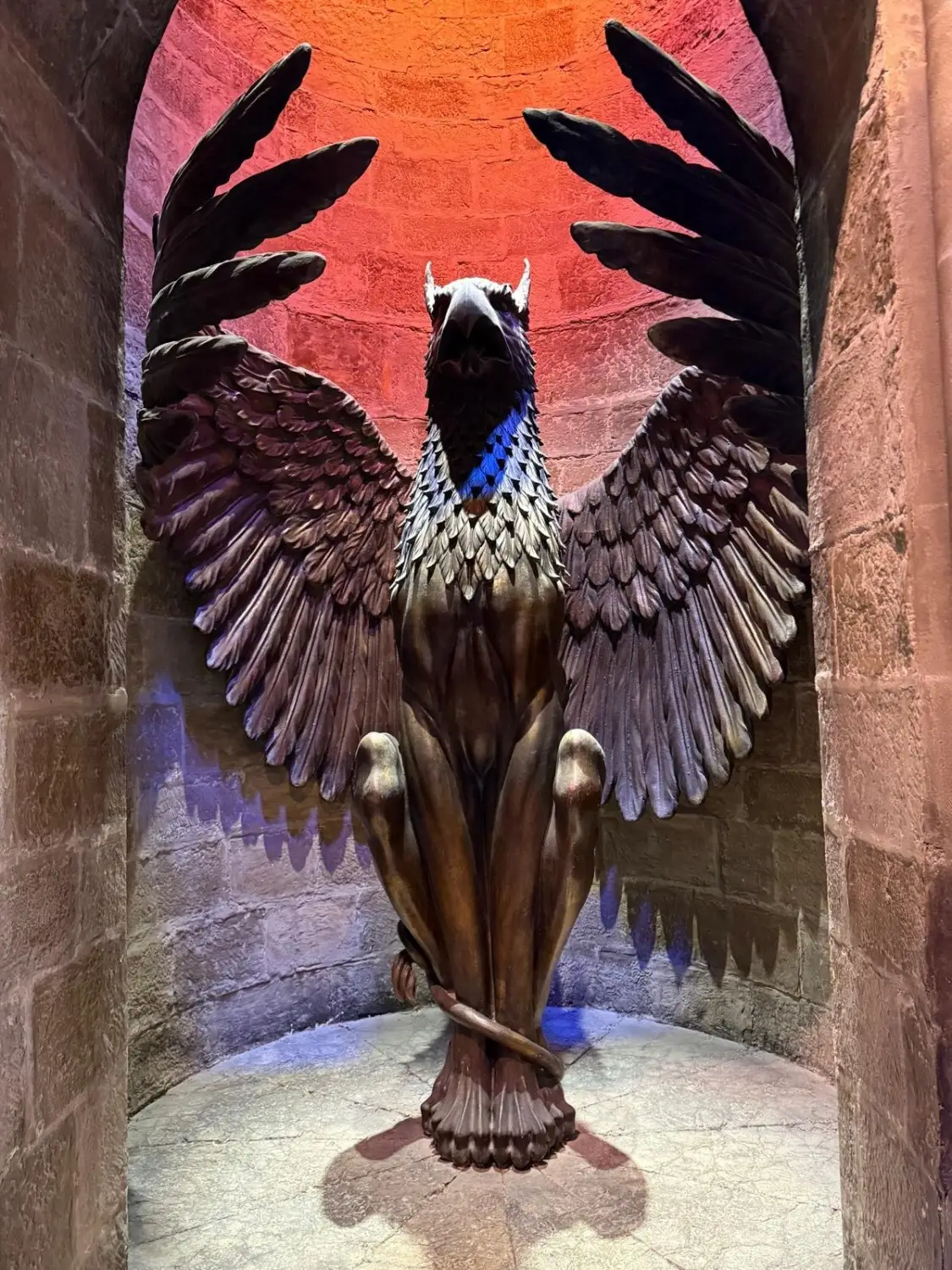 A large winged griffin statue with its wings spread wide, positioned inside a stone alcove with warm orange and purple lighting highlighting the textured surface.