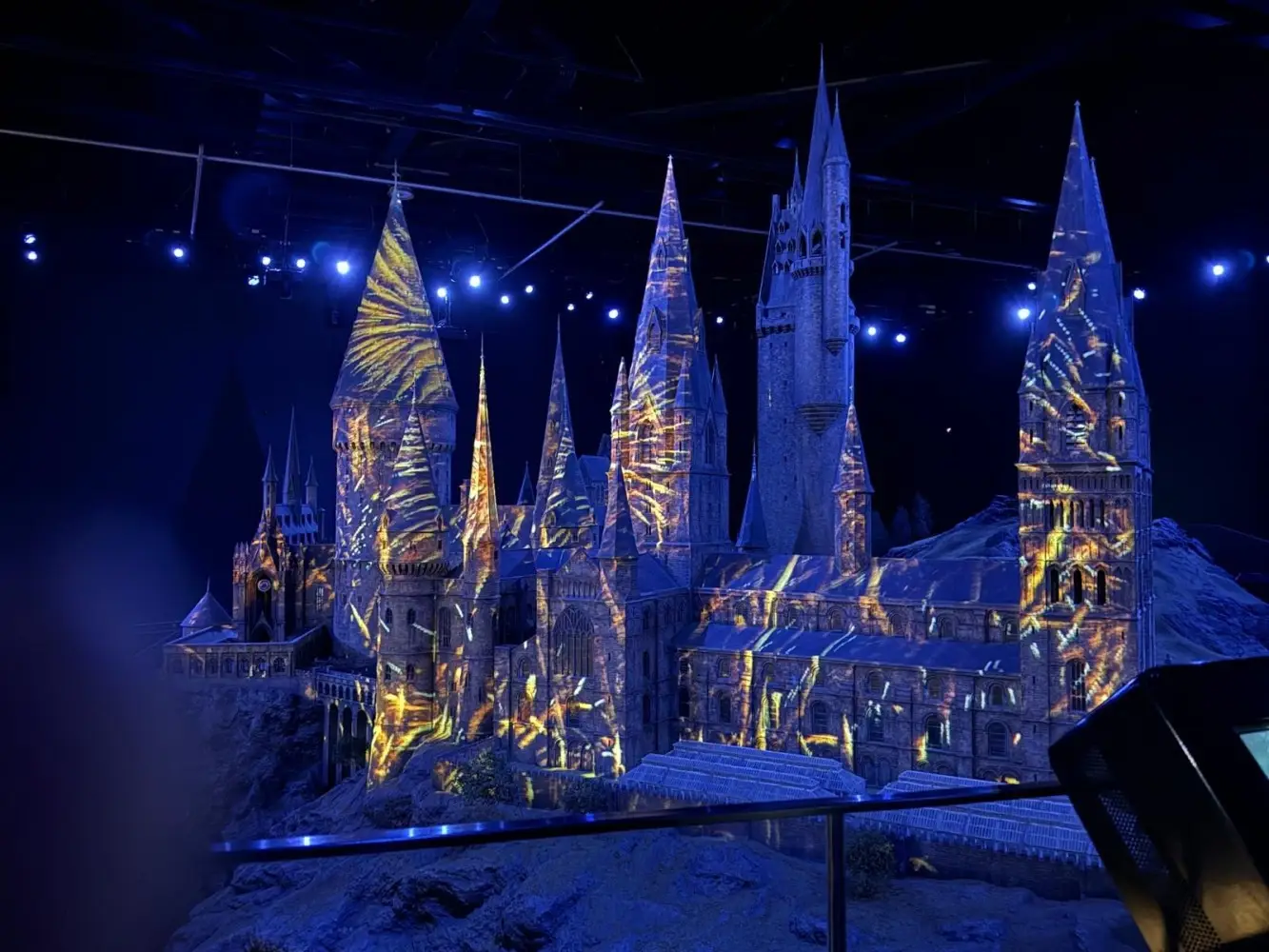 A detailed scale model of Hogwarts Castle lit with blue and golden projection effects, highlighting the towers and architecture in a dark exhibition room.