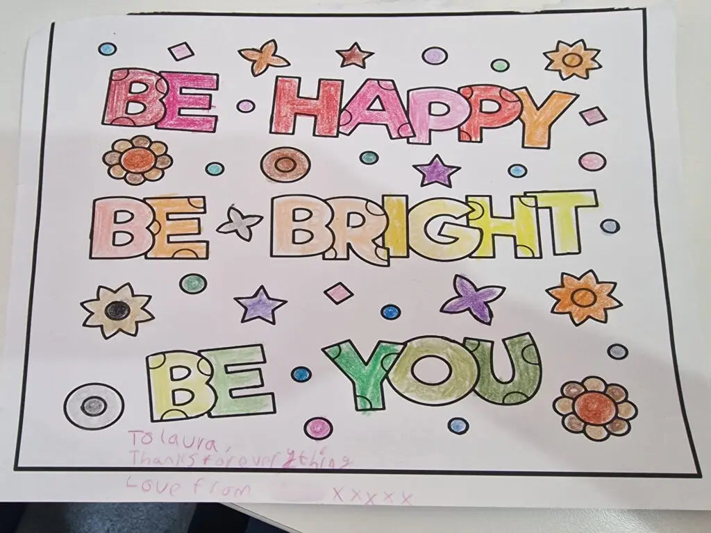Hand-drawn poster with colorful block letters reading “Be Happy, Be Bright, Be You.” The words are decorated with multicolored patterns and surrounded by small flowers, stars, dots, and geometric shapes. A thin black border frames the page, and a handwritten note appears at the bottom.