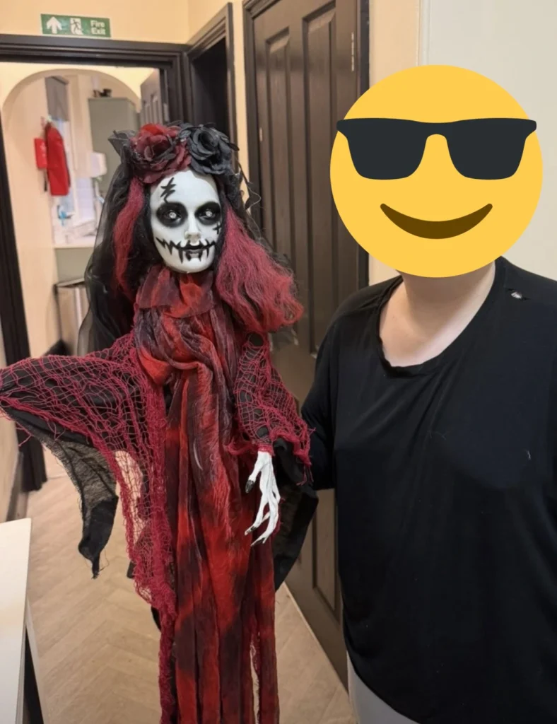 A young person stands beside a red-and-black Halloween hanging decoration featuring a spooky face, dark veil, and tattered fabric, displayed indoors as part of seasonal decorations.