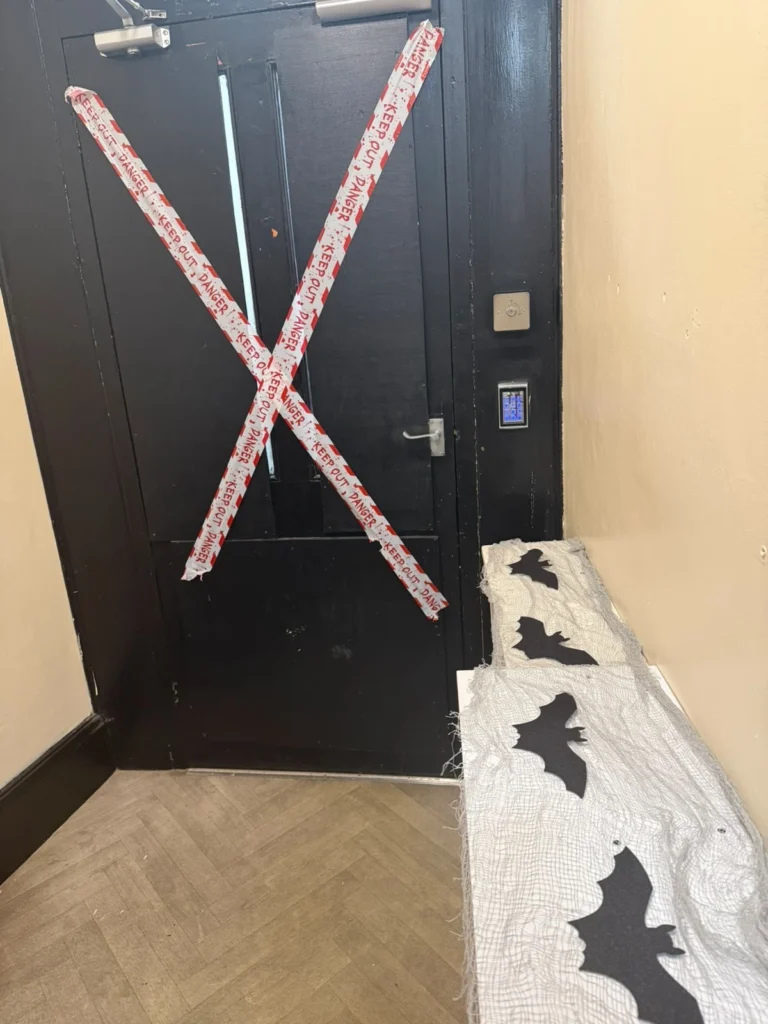 Black door decorated with Halloween-themed "Keep Out" warning tape arranged in an X shape, next to a display covered in white web fabric and black bat decorations.